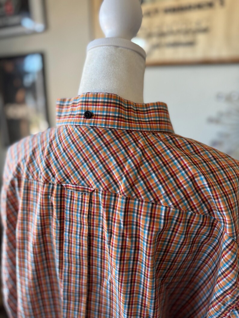 Puede incluir: Primer plano de una camisa abotonada con un estampado de cuadros en naranja, rojo, azul y blanco. La camisa se muestra en un maniqu&iacute;. El cuello tiene un solo bot&oacute;n negro.