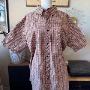 Puede incluir: Camisa abotonada de manga corta con un estampado a cuadros en naranja, rojo, azul y blanco. La camisa tiene cuello, bolsillo en el pecho y botones negros. La camisa se exhibe en un maniqu&iacute; blanco.