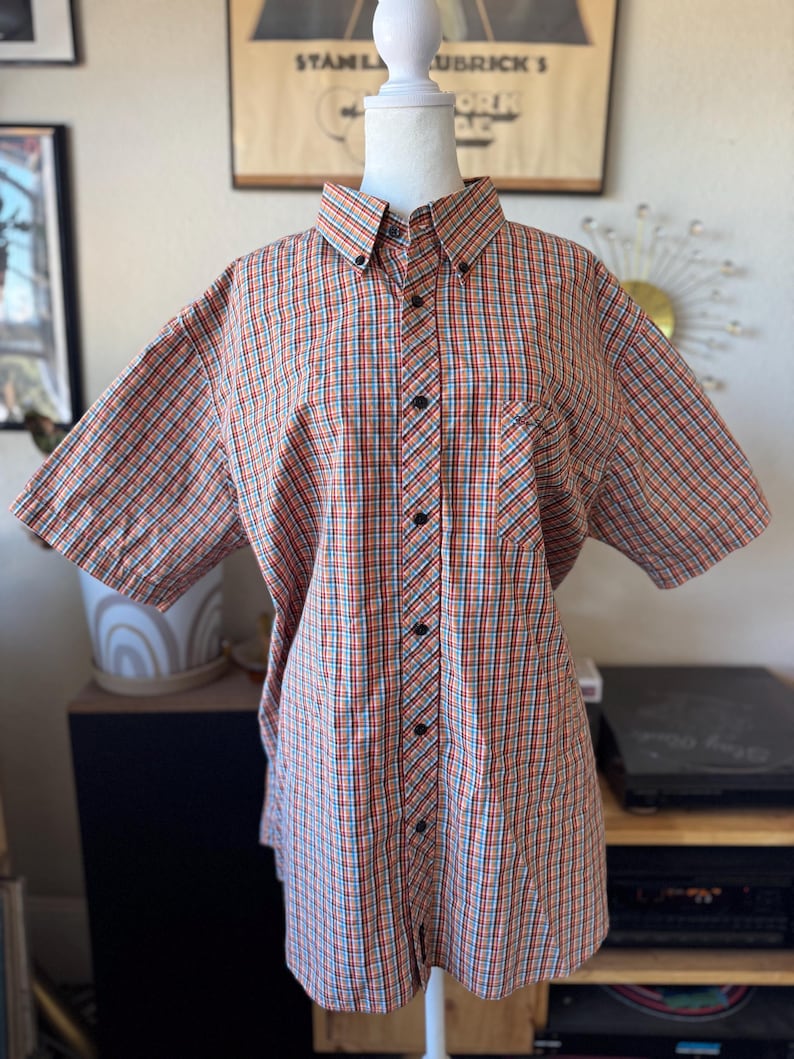 Puede incluir: Camisa abotonada de manga corta con un estampado a cuadros en naranja, rojo, azul y blanco. La camisa tiene cuello, bolsillo en el pecho y botones negros. La camisa se exhibe en un maniqu&iacute; blanco.