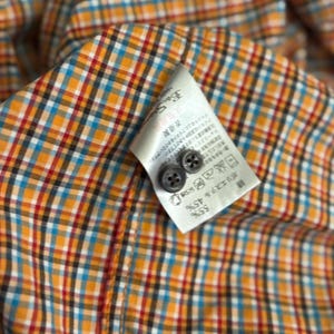 Puede incluir: Primer plano de una camisa a cuadros colorida con una etiqueta y dos botones. La camisa presenta un patr&oacute;n de cuadrados naranjas, azules, rojos y negros. La etiqueta tiene texto y dos peque&ntilde;os botones.