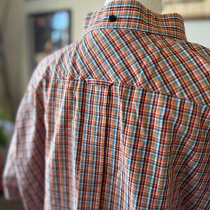 Puede incluir: Camisa abotonada de manga corta con un estampado a cuadros en naranja, verde azulado, rojo y blanco. La camisa se muestra en un maniqu&iacute;, mostrando la parte posterior del cuello y los hombros. Se ve un bot&oacute;n negro en la parte posterior del cuello.