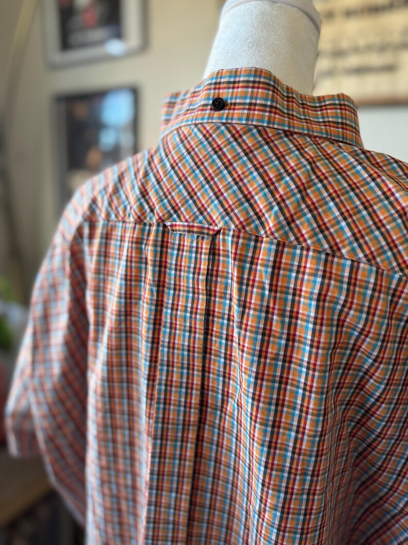 Puede incluir: Camisa abotonada de manga corta con un estampado a cuadros en naranja, verde azulado, rojo y blanco. La camisa se muestra en un maniqu&iacute;, mostrando la parte posterior del cuello y los hombros. Se ve un bot&oacute;n negro en la parte posterior del cuello.