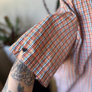 Puede incluir: Camisa de manga corta a cuadros naranja, azul y blanco. El pu&ntilde;o est&aacute; abotonado. La tela presenta un dise&ntilde;o de cuadros cl&aacute;sico, adecuado para climas c&aacute;lidos. Estilo casual.