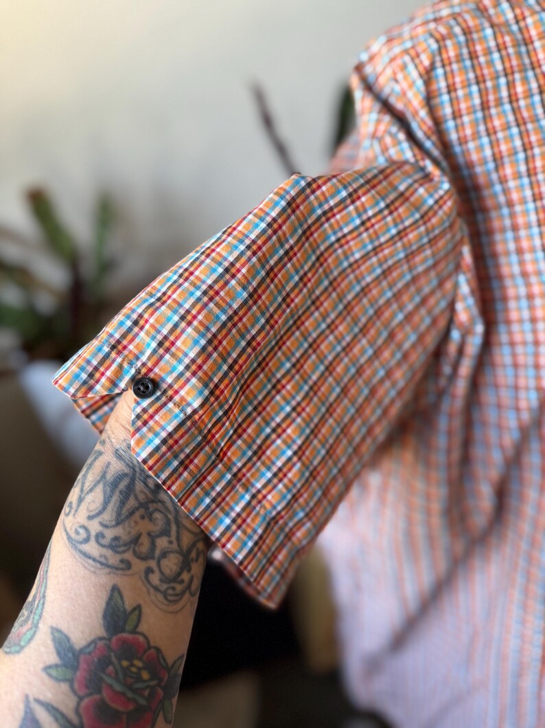 Puede incluir: Camisa de manga corta a cuadros naranja, azul y blanco. El pu&ntilde;o est&aacute; abotonado. La tela presenta un dise&ntilde;o de cuadros cl&aacute;sico, adecuado para climas c&aacute;lidos. Estilo casual.