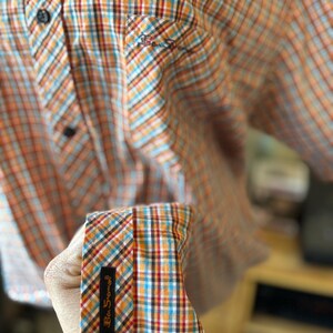 Puede incluir: Camisa abotonada a cuadros naranja, azul y blanco. La camisa tiene un peque&ntilde;o bolsillo en el pecho y un pu&ntilde;o con una etiqueta negra que dice "Ben Sherman". Se ven botones negros.