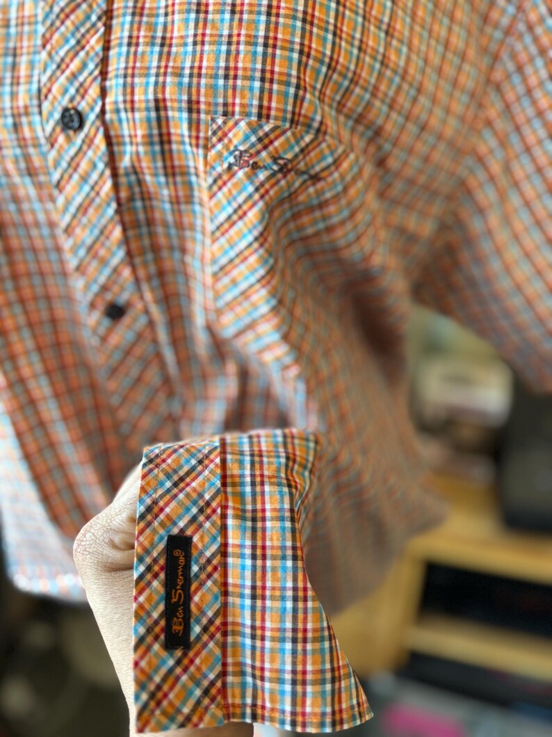 Puede incluir: Camisa abotonada a cuadros naranja, azul y blanco. La camisa tiene un peque&ntilde;o bolsillo en el pecho y un pu&ntilde;o con una etiqueta negra que dice "Ben Sherman". Se ven botones negros.