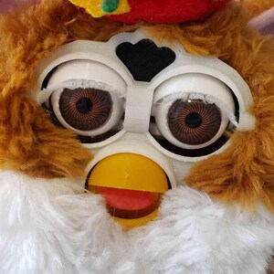 May include: A brown and white furry toy with a red and yellow hat. The toy has large, round eyes with black pupils and a yellow beak. The toy is wearing a red hat with a green and yellow bow.