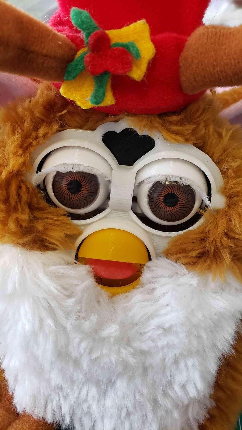 May include: A brown and white furry toy with a red and yellow hat. The toy has large, round eyes with black pupils and a yellow beak. The toy is wearing a red hat with a green and yellow bow.