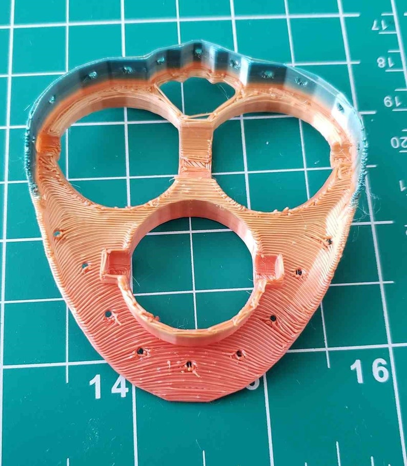 May include: A 3D printed orange and teal plastic part with three circular openings. The part has a textured surface and is designed with a rounded shape.