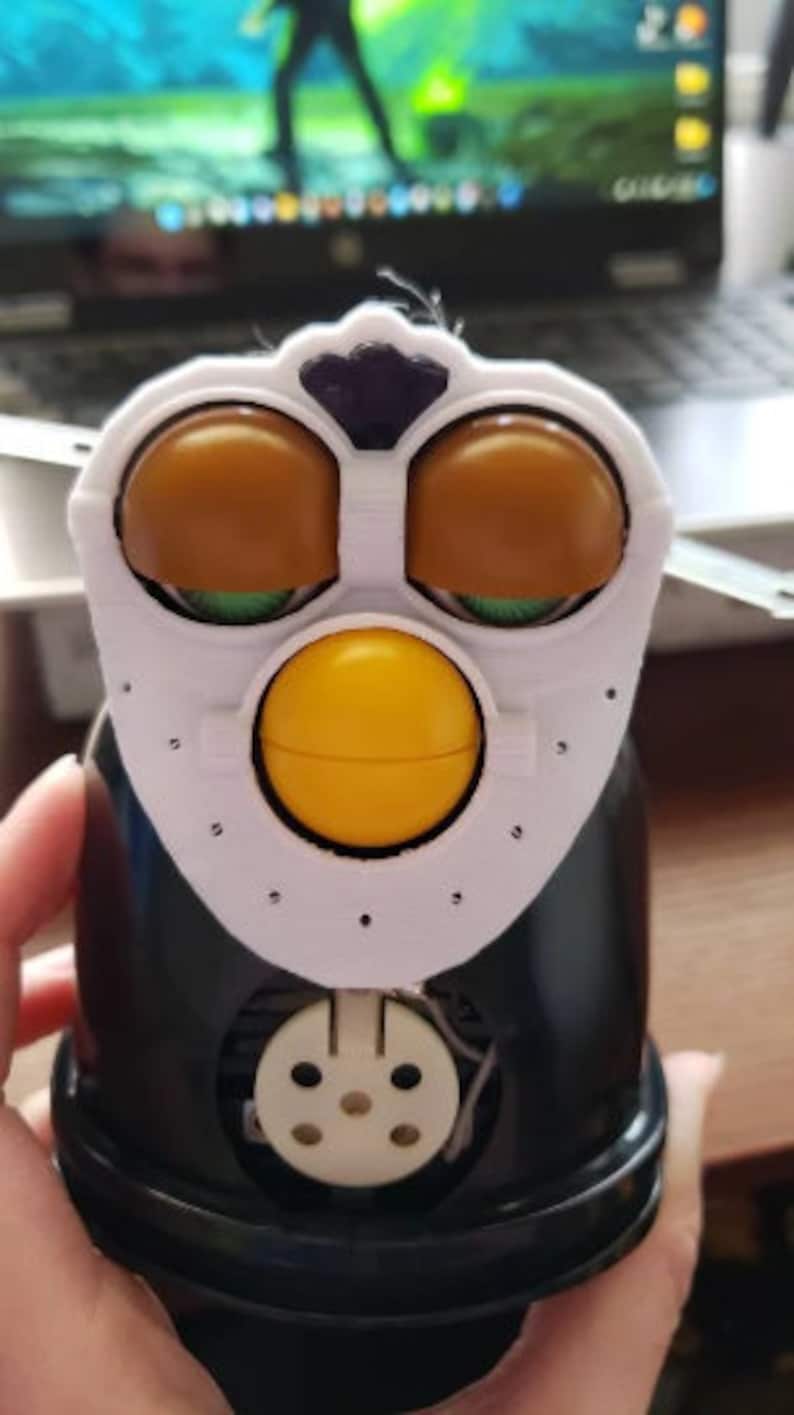 May include: A black and white toy penguin with brown eyes, a yellow beak, and a white face mask. The toy has a round body and a small, round head. The toy is made of plastic and has a hole in the bottom for batteries.