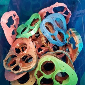 May include: A collection of 3D printed plastic heart-shaped molds in various colors, including green, blue, orange, and pink. The molds have multiple holes and are stacked on top of each other.