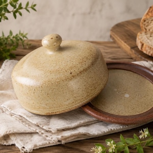 May include: A ceramic butter dish with a speckled cream-coloured dome lid featuring a small knob handle and a flat, dark brown circular base. The dish is crafted from clay with a visible texture and sits on a folded off-white fabric cloth.