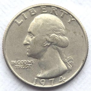 Puede incluir: Una moneda de cuarto de dólar estadounidense de plata de 1974. La moneda presenta un perfil de George Washington, con las palabras "LIBERTY" e "IN GOD WE TRUST" visibles. El año "1974" también se muestra en la moneda.
