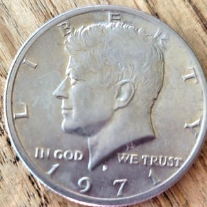 May include: A silver 1971 Kennedy half dollar coin. The obverse features a profile of John F. Kennedy, with the inscriptions "LIBERTY", "IN GOD WE TRUST", and the date "1971". The coin is on a wooden surface.