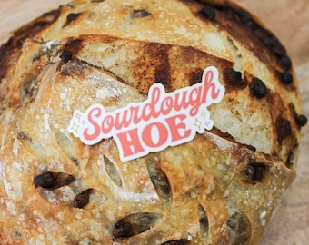 May include: Close-up of a loaf of sourdough bread with a sticker that reads "Sourdough HOE" in pink and red. The bread has a golden-brown crust with dark brown spots and decorative cuts.