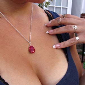 Puede incluir: Un collar de cadena de plata con un colgante de piedra roja. El colgante tiene forma ovalada y un corte facetado. El collar lo lleva una persona con una camisa azul.