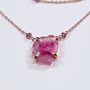 May include: A delicate rose gold necklace with a raw pink tourmaline pendant and small pink tourmaline accents. The necklace is layered with two chains.