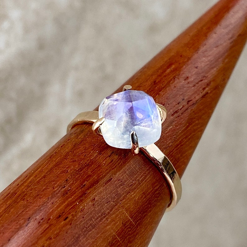 K&ouml;nnte beinhalten: Ein goldener Ring mit einem gro&szlig;en, schillernden Mondstein, der in einer Zargenfassung sitzt. Der Stein ist hellblau mit einem Hauch von Lila.