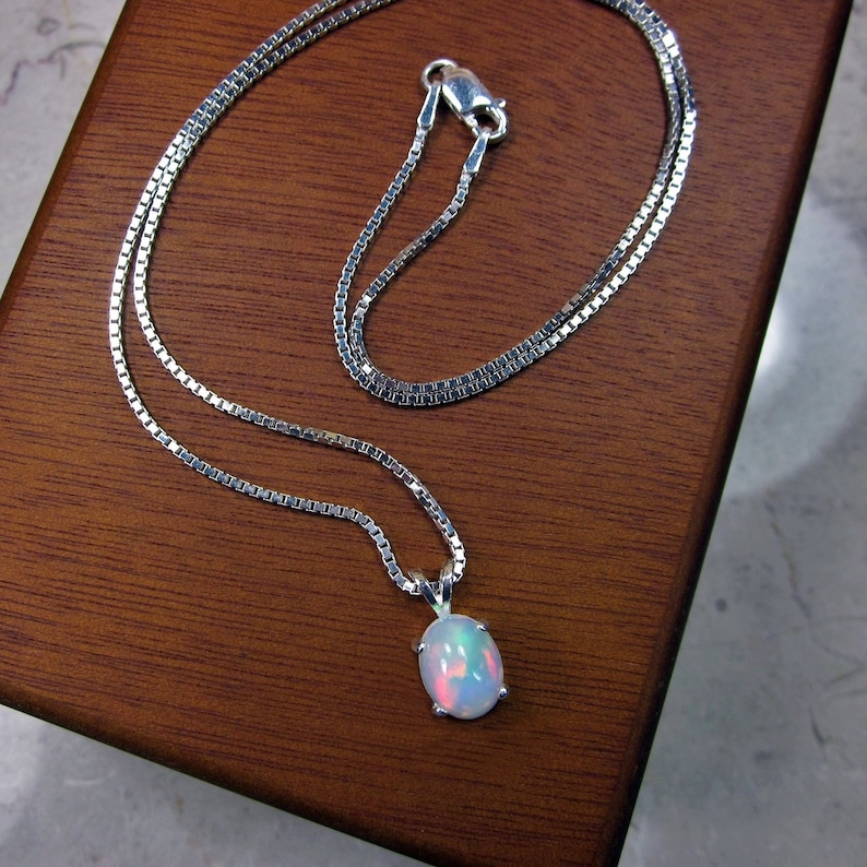 K&ouml;nnte beinhalten: Eine silberne Halskette mit einem kleinen, ovalen Opalanh&auml;nger. Der Opal zeigt ein Farbspiel mit rosa, blauen und gr&uuml;nen Farbt&ouml;nen. Die Kette ist eine feine Boxkette, und der Anh&auml;nger ist in einer silbernen L&uuml;nette gefasst.