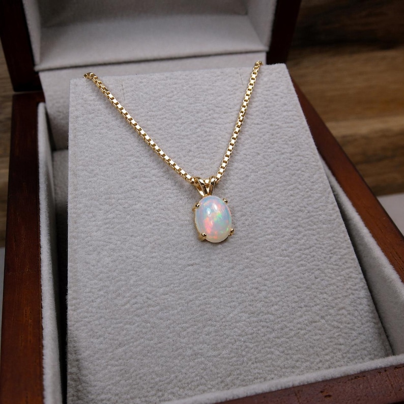 K&ouml;nnte beinhalten: Eine goldene Halskette mit einem ovalen Opalanh&auml;nger, pr&auml;sentiert in einer Holzschatulle. Der Opal hat eine milchig-wei&szlig;e Basis mit Farbflecken. Die Kette ist eine filigrane Box-Kette.