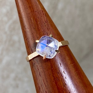 K&ouml;nnte beinhalten: Ein goldener Ring mit einem klaren, schillernden Mondstein, der in einer quadratischen Form gefasst ist. Der Ring befindet sich auf einem Holzst&auml;nder.