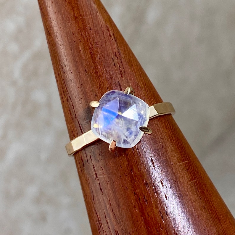 K&ouml;nnte beinhalten: Ein goldener Ring mit einem klaren, schillernden Mondstein, der in einer quadratischen Form gefasst ist. Der Ring befindet sich auf einem Holzst&auml;nder.