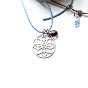 Op de afbeelding: Een zilveren ketting met een delicate, ronde mandala-hanger. De hanger is bevestigd aan een lichtblauw koord en heeft een kleine, paarse glazen kraal.