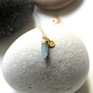 Könnte beinhalten: Eine zarte goldene Halskette mit einem hellblauen Kristallanhänger und goldenen Akzenten. Die Halskette enthält einen kleinen, runden Goldanhänger. Der Schmuck wird auf einem hellgrauen Stein präsentiert, was eine minimalistische Ästhetik schafft.