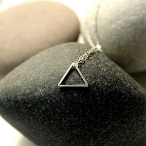 Könnte beinhalten: Eine silberfarbene Halskette mit einem geometrischen Dreiecksanhänger. Der Anhänger hängt an einer filigranen Kette. Die Halskette wird auf einem dunkelgrauen Stein präsentiert, mit anderen Steinen im Hintergrund.