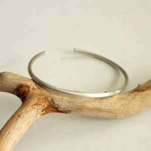 Könnte beinhalten: Ein schlichtes silbernes Armreif-Armband mit glatter, abgerundeter Oberfläche. Das Armband wird auf einem Stück Treibholz präsentiert.
