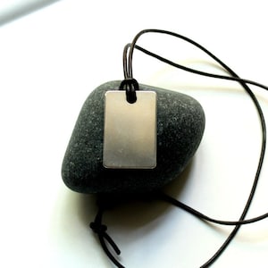 Puede incluir: Un colgante rectangular de plata con un agujero en la parte superior cuelga de un cordón de cuero marrón. El colgante está apoyado en una roca gris.