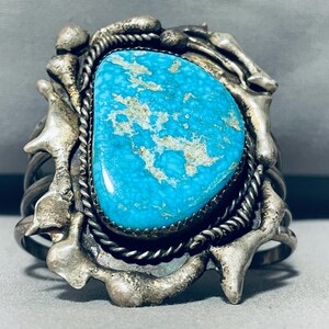 Puede incluir: Un brazalete de plata y turquesa con una piedra turquesa grande e irregular engastada en un engaste de plata. El brazalete tiene un diseño único y orgánico con un detalle de alambre de plata trenzado.