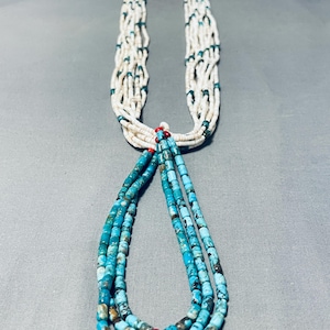 Puede incluir: Un collar de múltiples hilos con cuentas de turquesa y blancas. El collar tiene una sección larga y colgante con cuentas de turquesa y algunas cuentas de coral rojo en la parte inferior.