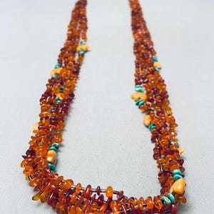 May include: A multi-strand necklace featuring numerous small, polished amber chips in varying shades of orange and brown, interspersed with small, smooth, light-colored stone beads and tiny, vibrant turquoise-colored accent pieces.