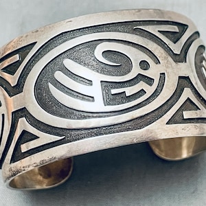 May include: A wide silver cuff bracelet featuring a polished finish with recessed, oxidized dark areas. The surface displays intricate, stylized geometric and abstract curved patterns that wrap around the exterior of the metal band.