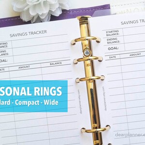 May include: A white planner page with a savings tracker table. The table has columns for date, amount, and balance. The planner is bound with gold rings. The text "SAVINGS TRACKER" is at the top of the page.