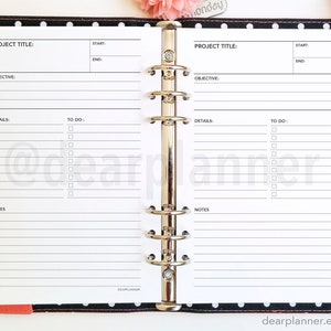 K&ouml;nnte beinhalten: Ein wei&szlig;er Planer mit schwarzen Linien und einem schwarz-wei&szlig;en Polka-Dot-Umschlag. Der Planer hat einen Metallringbinder und zwei Seiten, die zu einer Projektplanungsvorlage ge&ouml;ffnet sind. Die Vorlage hat Abschnitte f&uuml;r Projekttitel, Ziel, Details, zu erledigende Aufgaben und Notizen.