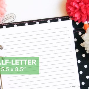 May include: A black and white polka dot binder with a white page open inside. The page is lined and has the text "HALF-LETTER 5.5 x 8.5" printed on it. The binder has a silver ring mechanism and a silver clip on the side. There are two pink and white pom-poms on the top and right side of the binder.