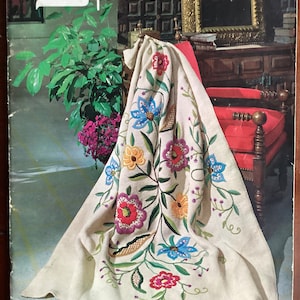 May include: A vintage magazine cover featuring a white embroidered throw blanket with colorful floral designs. The magazine is titled "Spinnerin" and the text "Fashions in Living" is printed on the bottom right corner. The magazine is from Volume 168 and the price is $1.25.