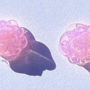 May include: Two pink, flower-shaped glass objects with a translucent appearance are displayed. The objects cast dark shadows on a light-colored surface. The glass has a bubbly texture, and the overall aesthetic is delicate and decorative.