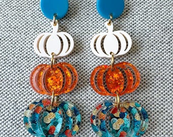 May include: A pair of dangle earrings featuring a solid blue circular stud at the top, followed by a white pumpkin, an orange glitter pumpkin, and a blue and multicolored glitter pumpkin, all connected by small gold-toned metal jump rings.