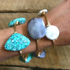 Puede incluir: Tres pulseras de alambre dorado envueltas con piedras de diferentes colores. La primera pulsera tiene una piedra turquesa, la segunda una piedra gris y la tercera una piedra blanca.