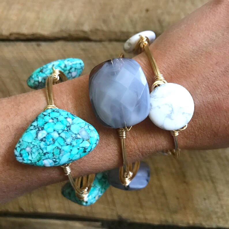 Puede incluir: Tres pulseras de alambre dorado envueltas con piedras de diferentes colores. La primera pulsera tiene una piedra turquesa, la segunda una piedra gris y la tercera una piedra blanca.