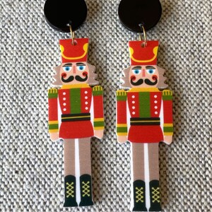 May include: A pair of Christmas-themed earrings featuring a nutcracker design. The earrings are made of wood and have a black circle at the top. The nutcracker is wearing a red and green uniform with a gold belt and black boots.