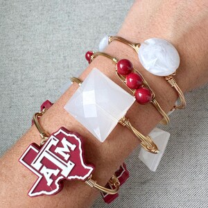 May include: A collection of gold wire bracelets adorned with white, red, and maroon accents. One bracelet features a maroon Texas-shaped charm with "ATM" in white. Other bracelets have white geometric shapes and red beads.