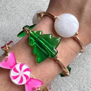 May include: Three gold wire bracelets with different charms. The first bracelet has a white round charm, the second has a green Christmas tree charm, and the third has a pink and white candy cane charm.