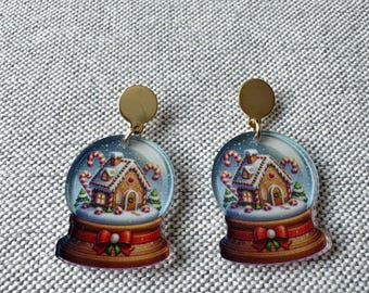 May include: A pair of dangle earrings featuring a snow globe design. Each earring displays a gingerbread house inside a clear dome with candy canes, set upon a brown base decorated with a red ribbon and a green bow, attached to a gold stud.