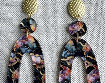 Op de afbeelding: Een paar statement oorbellen met een goudkleurige, getextureerde stud, een kleine ronde hanger en een grote boogvorm. De oorbellen hebben een gemarmerd ontwerp met zwarte, gouden, paarse en oranje tinten.