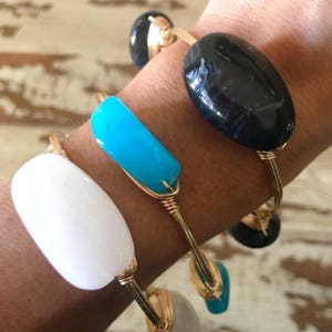 May include: Three gold wire wrapped bracelets with different colored stones. The bracelets feature a white, turquoise, and black stone.