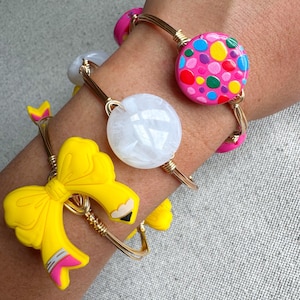Puede incluir: Tres pulseras de alambre dorado con diferentes dijes. La primera pulsera tiene un dije blanco texturizado. La segunda pulsera tiene un dije rosa con lunares de colores. La tercera pulsera tiene un dije de lazo amarillo.